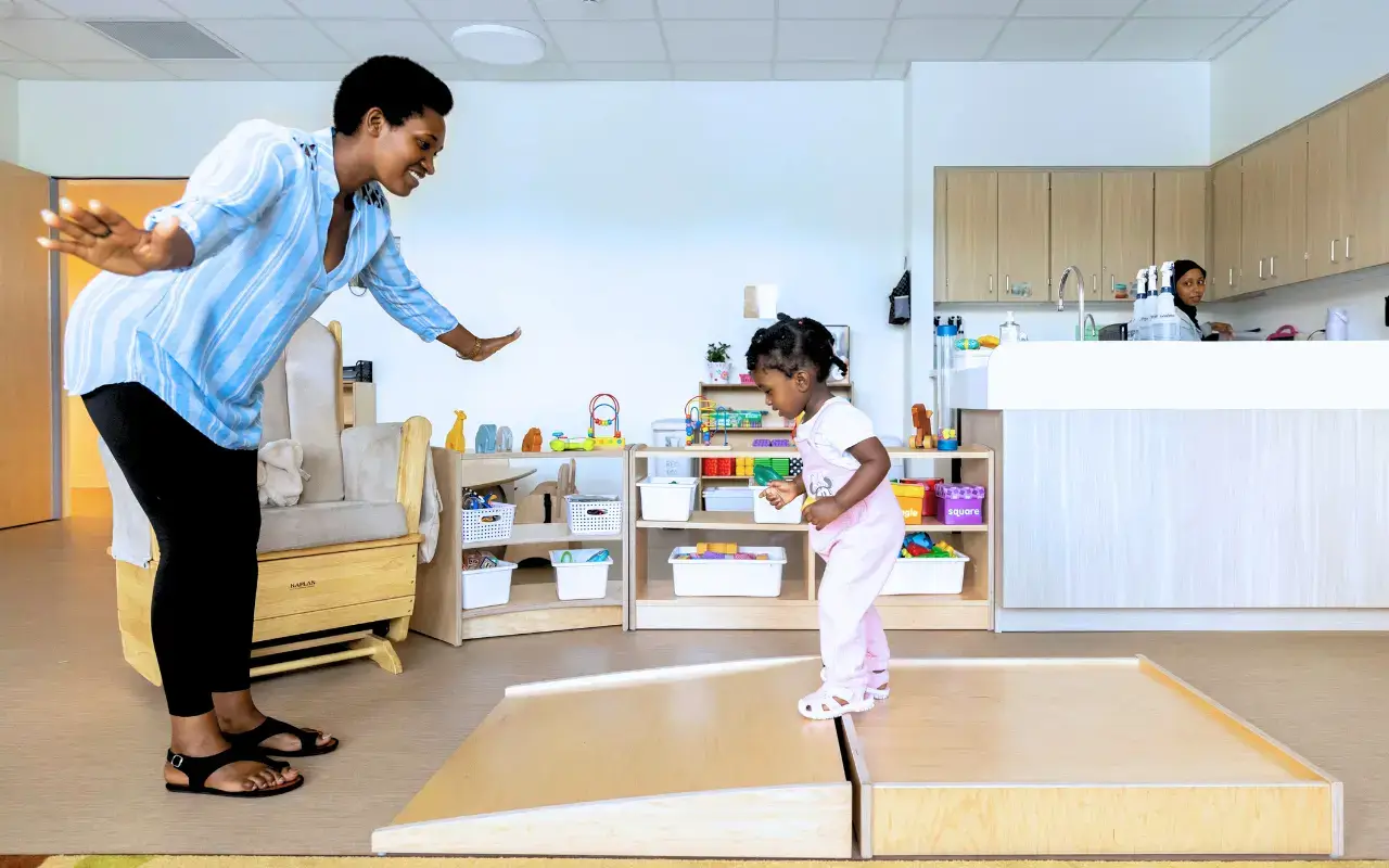 An educator encourages a child to walk up a Natural Pod Imagine Infant Ramp and Platform.