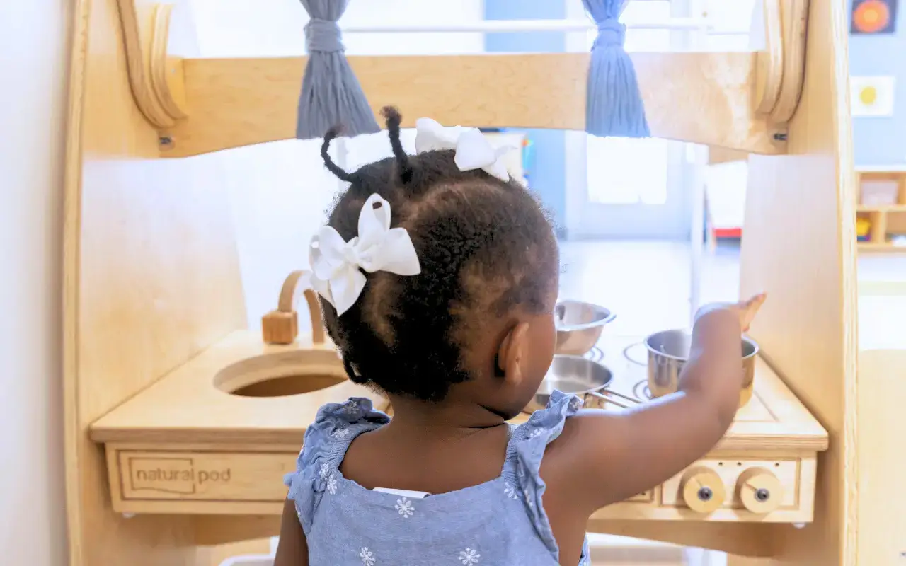 A toddler with white bows in her hair plays with a Natural Pod Imagine Play Kitchen.