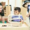 A teacher smiles at a young learner playing with a wooden car track at Tiny Scholars
