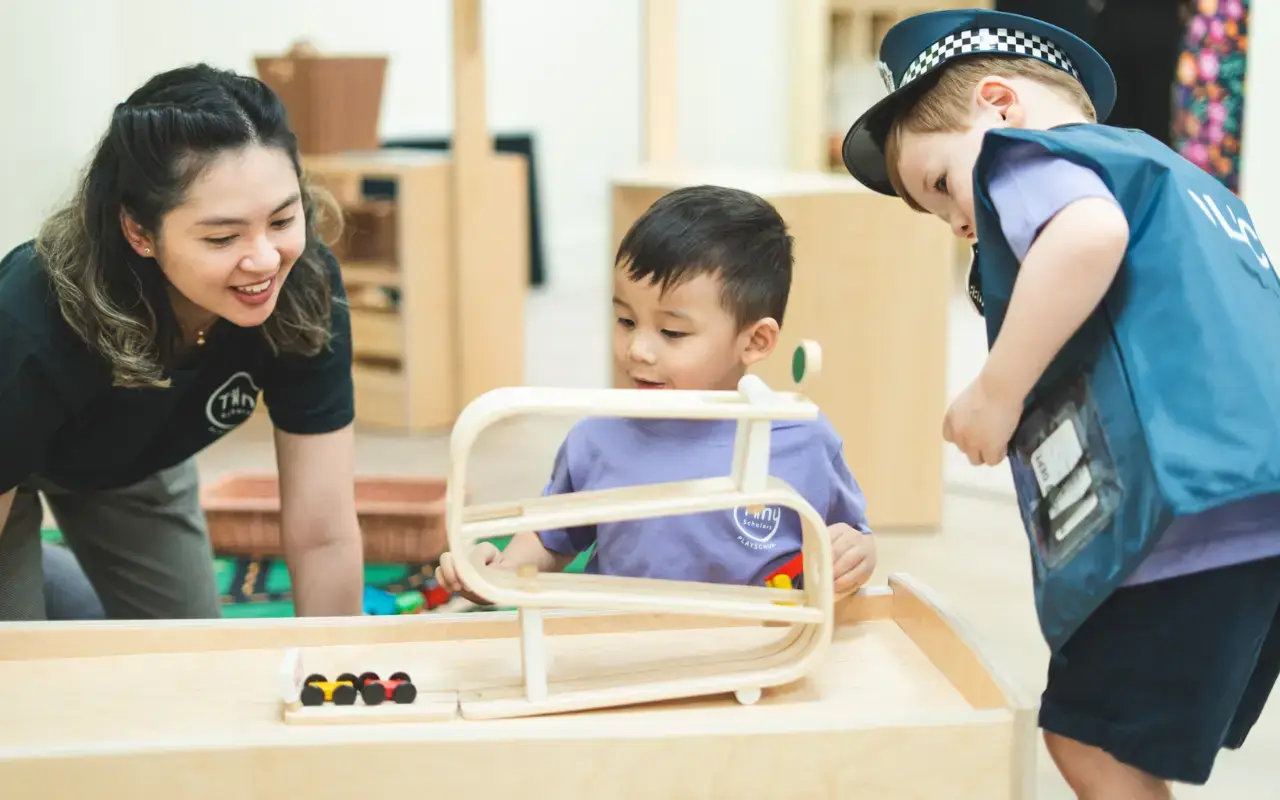 A teacher smiles at a young learner playing with a wooden car track at Tiny Scholars