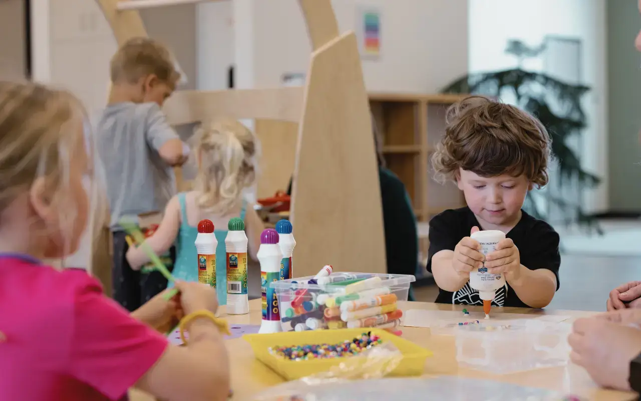 Close-up shot of a young child with curly hair concentrating while applying white glue to a piece of art at a Natural Pod Reach table in a brightly lit learning space. Other children and art supplies are visible in the foreground and background.