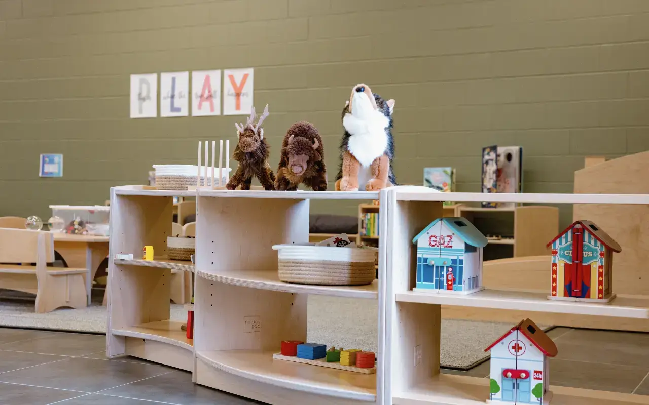 Natural Pod Evergreen shelves with rounded edges displays stuffed woodland animals and miniature playhouses in an early learning classroom. The background wall is painted olive green with the word "PLAY" spelled out.
