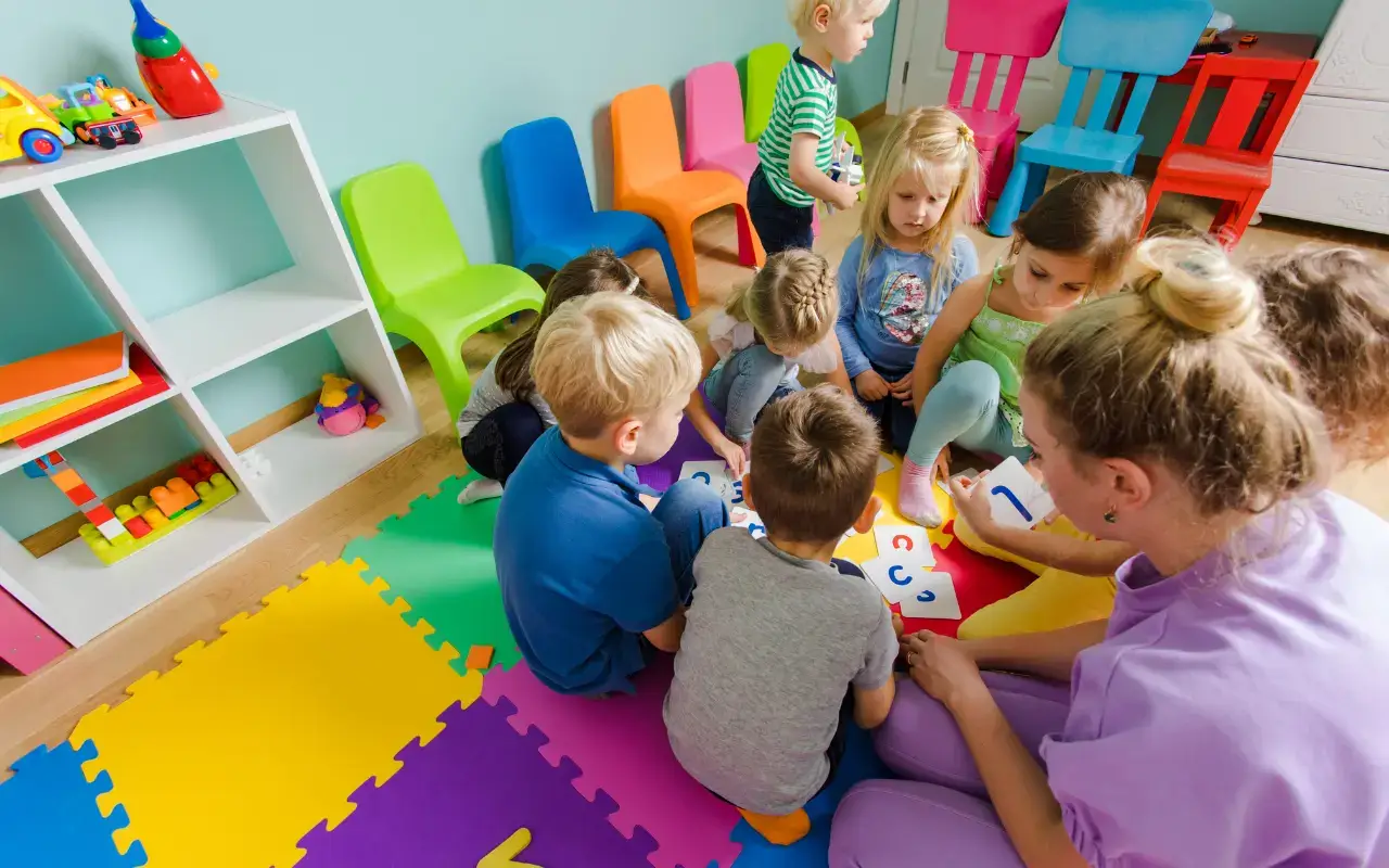A teacher and children sit on a colorful mat surrounded by bright plastic chairs and shelves, all examples of preschool furniture.