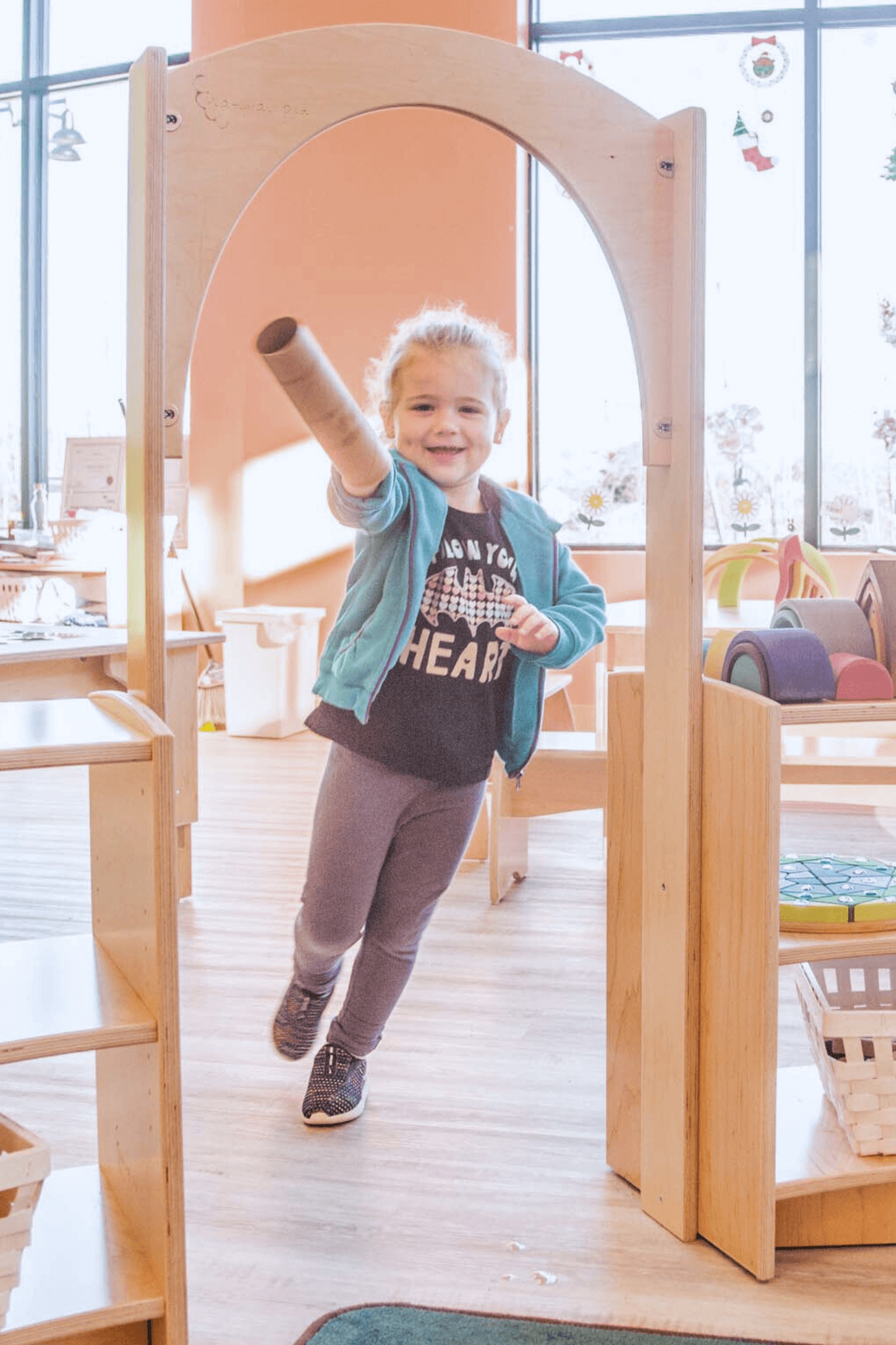 A smiling child runs through an Evergreen Archway, part of a Natural Pod preschool furniture set with shelving.