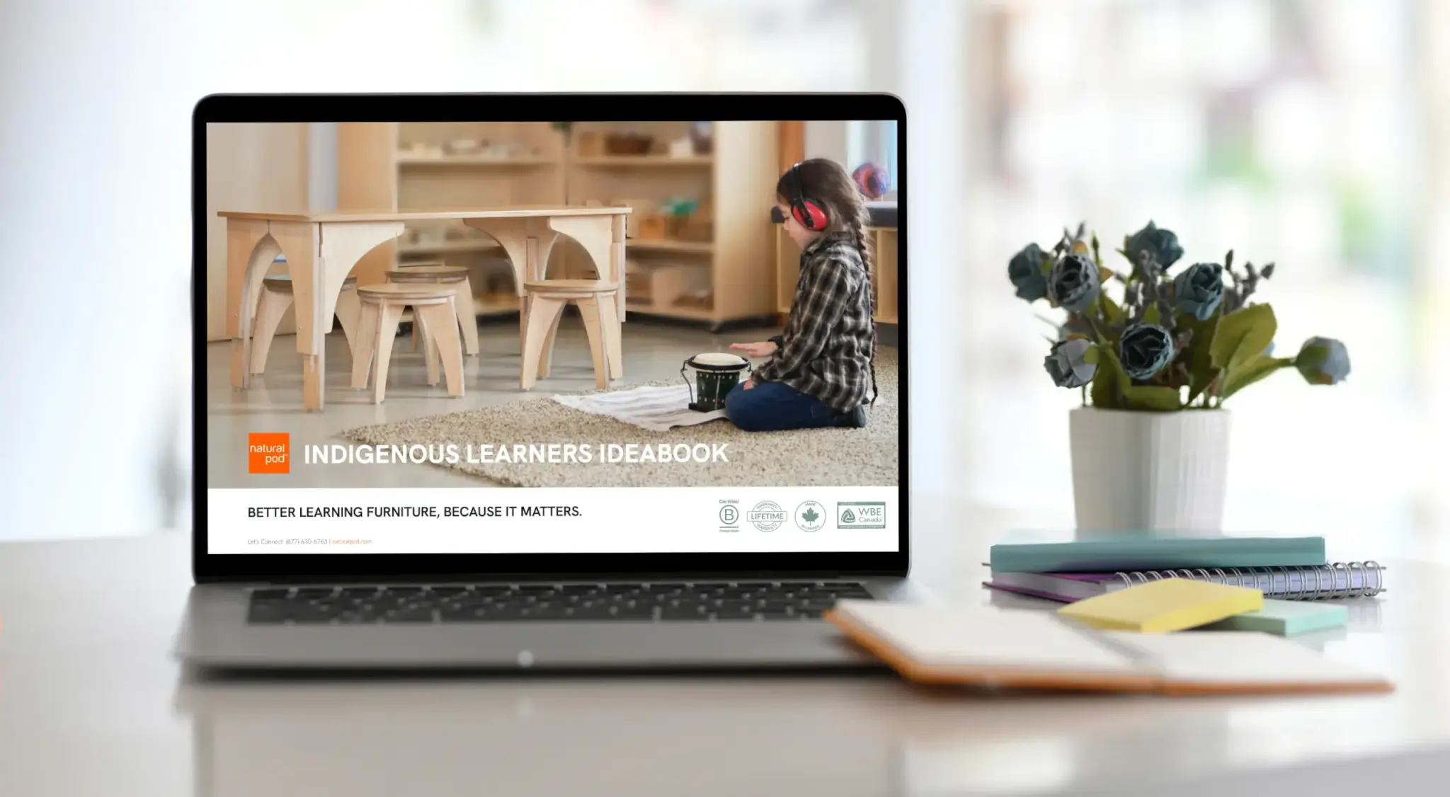 A laptop displays the Indigenous Learners Ideabook next to a notebook and plant