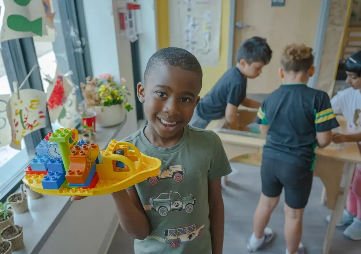 A student smiles with a toy boat as others play at a Reach Nature Table in the background of a St. Genevieve Catholic School classroom.