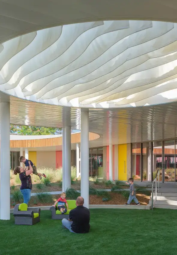 Families relaxing in a courtyard under wavy fabric shade sails.